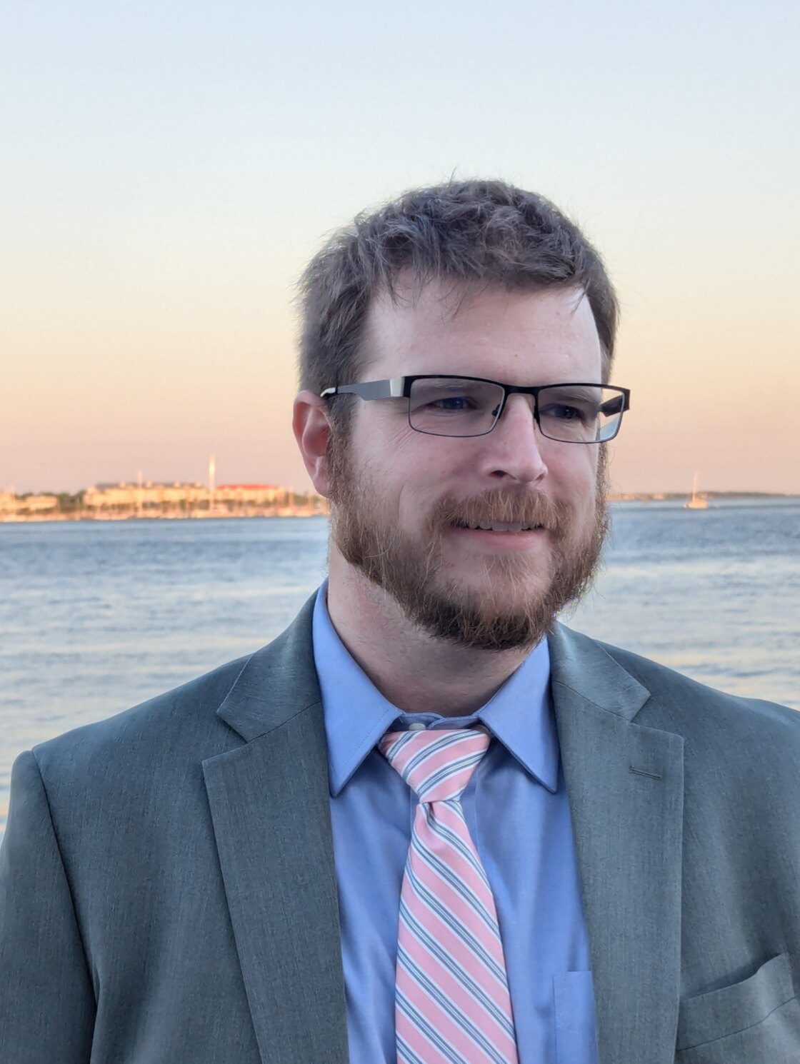Samuel Price standing by the water in a gray suit and pink striped tie at sunset.